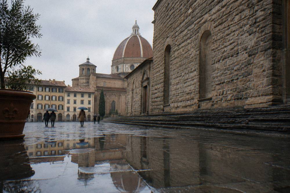 Seen low across rain-darkened paving toward the unfinished rough stone flank of San Lorenzo, a few umbrellas tilted far away in the square seen only from behind