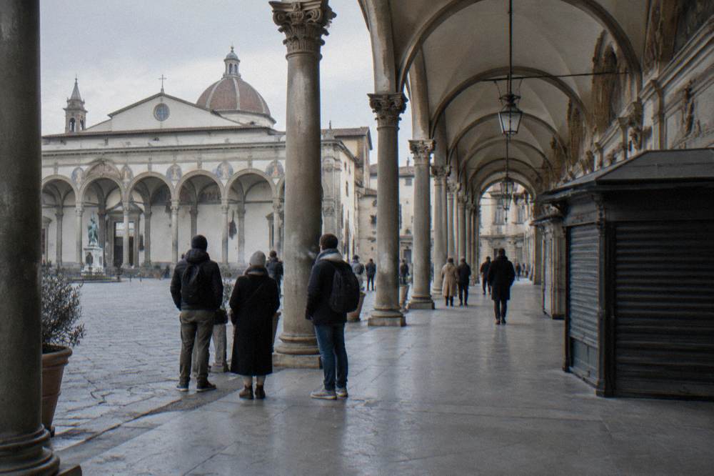 Viewed along the arched portico colonnade as newcomers gather on the smooth pruned-stone pavement facing the arcades and sculpted lunettes, breath fogging