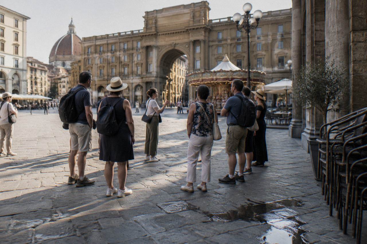 A loose ring of travellers assembled around a guide on the pale stone paving of Piazza della Repubblica, cafe chairs stacked nearby, last night's spilled coffee drying on the flagstones