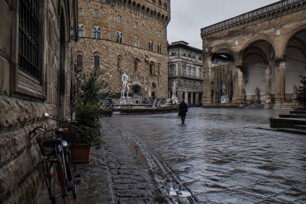 A cobbled lane running toward the square's pietra forte arcades, damp ruts between worn cobbles, a stray bicycle bell glinting, one distant figure from behind