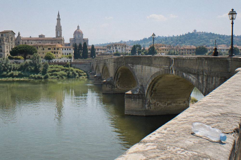 From the downstream parapet of Ponte alle Grazie looking along its stone arches over the green Arno, sun-bleached parapet, a crumpled water bottle left on the ledge