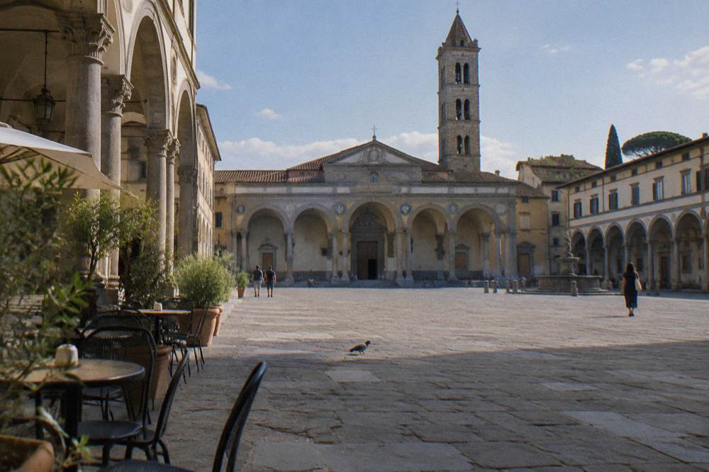 Across the symmetrical arcaded piazza toward its stone bell tower and grey pietra serena porticoes, scattered cafe chairs, a pigeon pecking between the paving joints