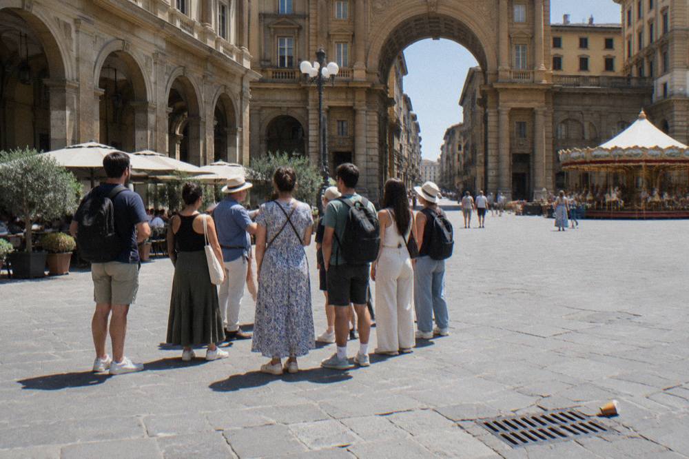 A walking group gathered from behind on the pale stone paving of the arcaded square, its tall arches and ochre facades in full sun, a dropped paper cup near a drain