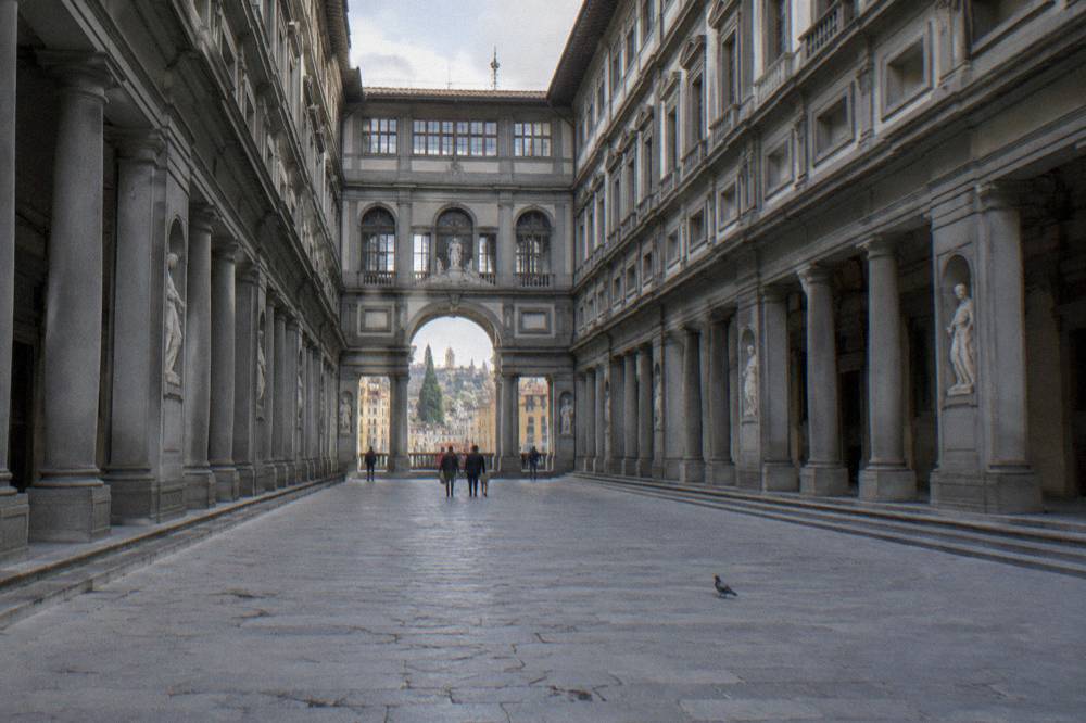 Looking along the long arcaded court of Piazzale degli Uffizi from its open end, grey pietra serena columns receding to a tall arch at the far end, pale flagstones