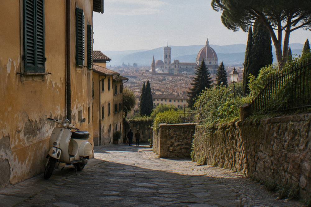 Low along a narrow cobbled lane below the Piazzale terraces, ochre plaster walls with flaking paint, closed green shutters, dry gravel and a parked Vespa casting shade