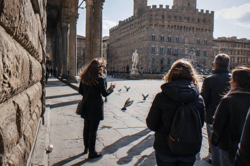 A guide seen from behind beside a rusticated stone wall and grey columned arcade, listeners' shadows long across worn trachyte paving, pigeons scattering, a dropped paper cup near the kerb