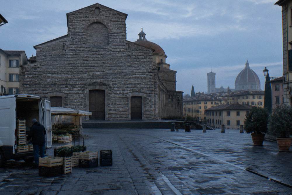 Looking across the empty square toward the church's rough unfinished stone facade, damp ruts between cobbles, a market van unloading crates, terracotta rooftops still dark against the haze