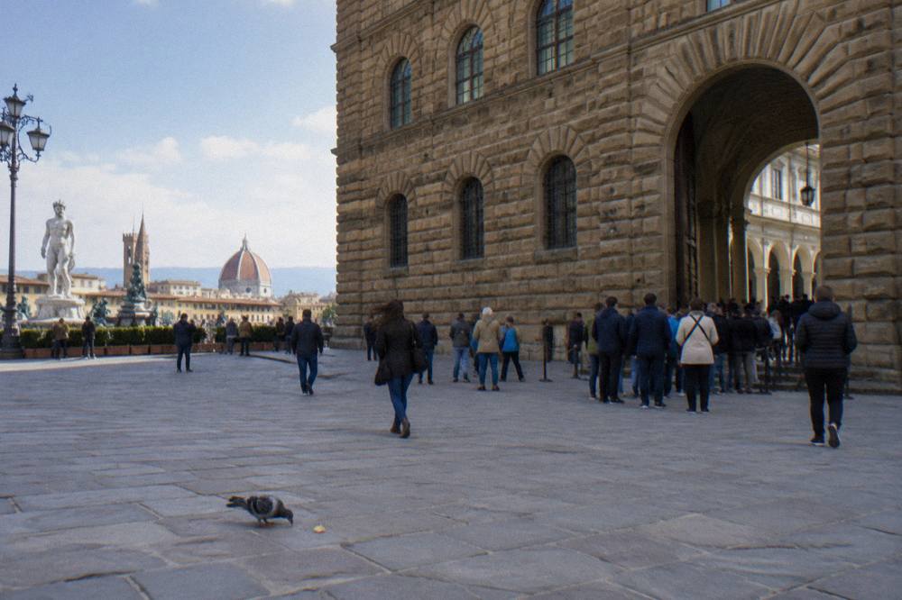 Seen across Piazza della Signoria toward the Uffizi's rusticated stone facade and arched courtyard entrance, tourists queuing in thin jackets