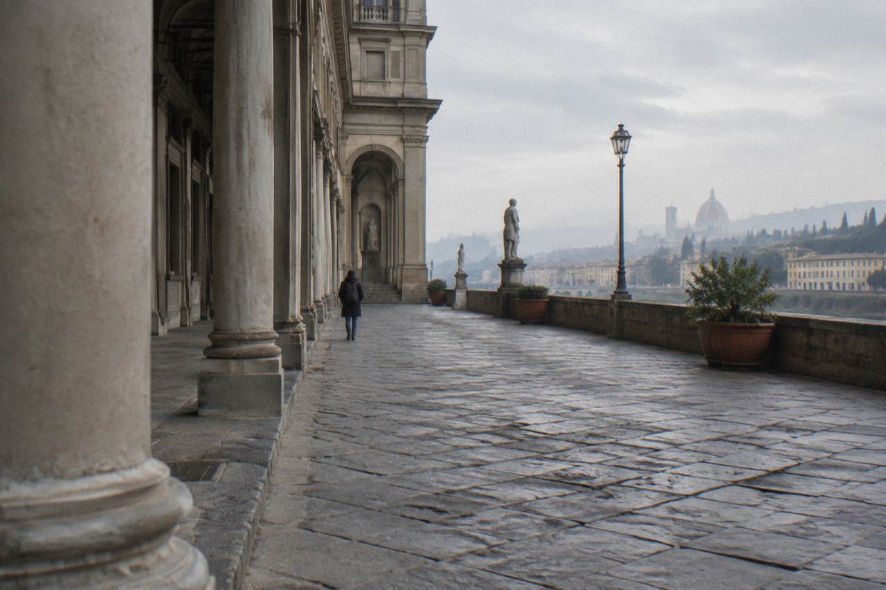 An off-centre low view along the arcade of Piazzale degli Uffizi, one pale stone column close in the left foreground, uneven paving, the far end of the court lost in haze