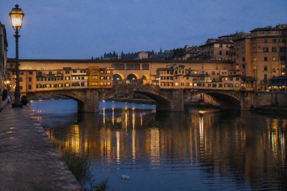 From a low riverside embankment across the Arno, the gold-lit arches and jutting shopfronts of the old bridge reflected in dark water, a stray plastic bottle drifting past the stone pier
