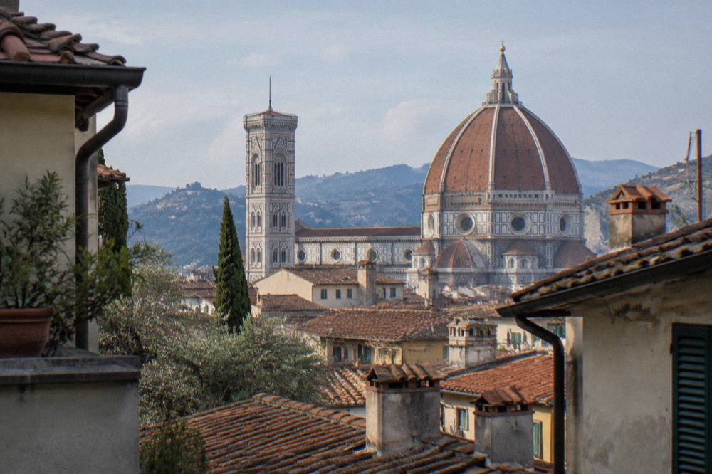 Viewed from a low rooftop gap between narrow terracotta-roofed houses, Brunelleschi's great tiled dome and Giotto's slim marble bell tower rising over dusty chimney stacks in pale light