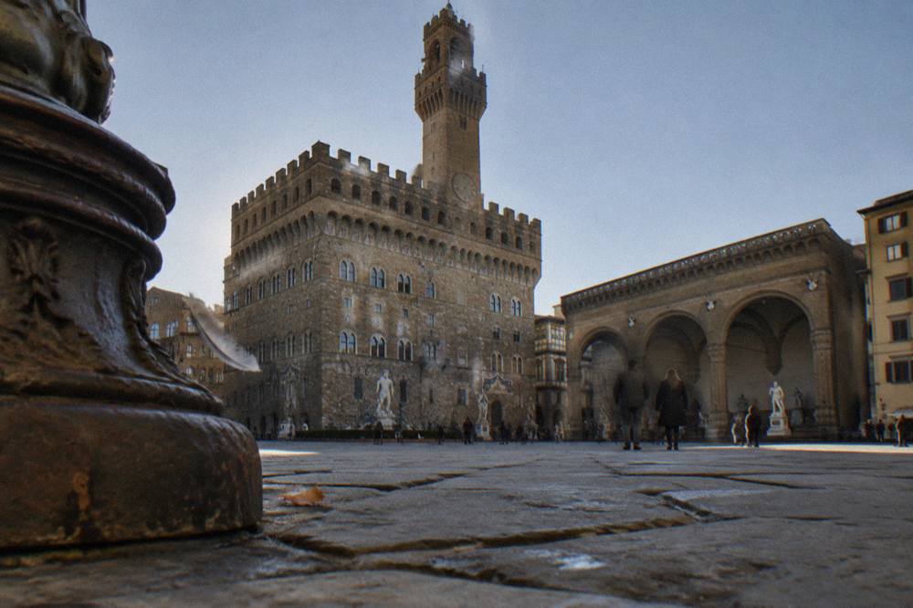 Low across worn pietra forte paving toward Palazzo Vecchio's crenellated tower and the open stone arches of the Loggia, clear sky, a stray pigeon feather drifting past a rusted lamp base