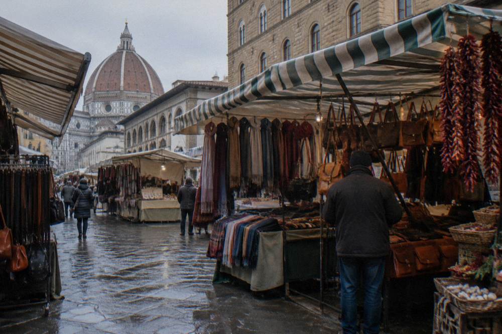 The open-air market stalls on the street around San Lorenzo, striped canvas awnings over tables of leather goods and scarves, strings of dried peppers hanging at one stall, wet paving