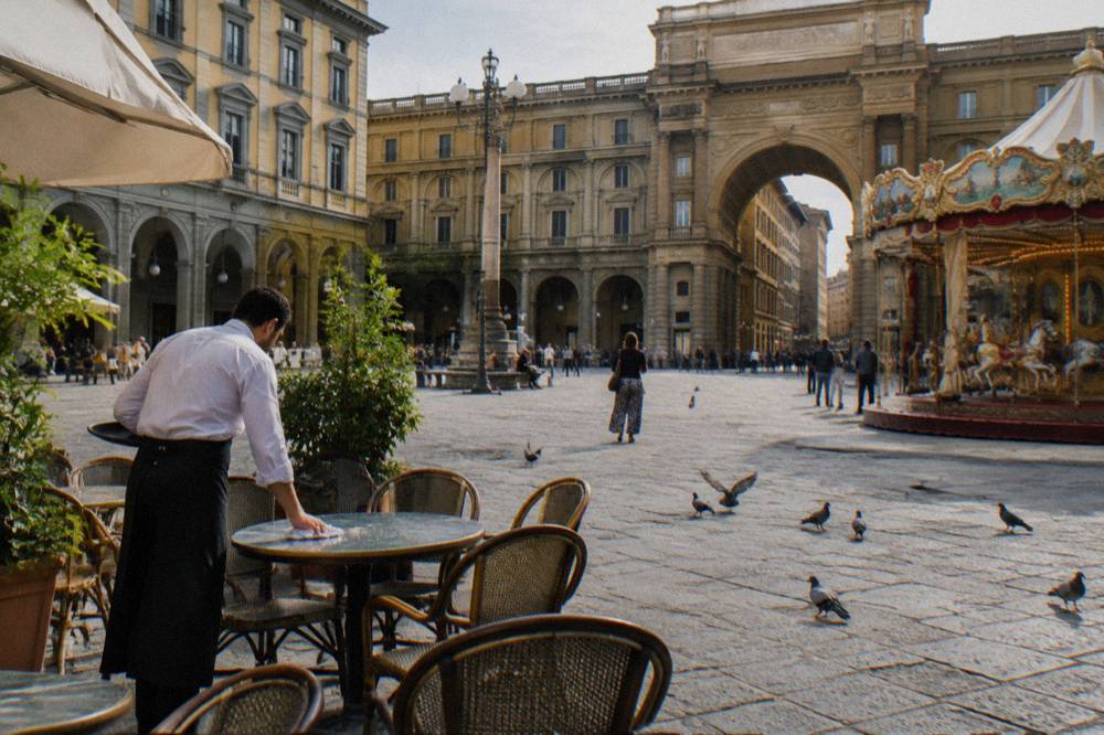 A wide public square ringed by ochre plaster palazzi and grey pietra serena arcades, a leafy carousel canopy at one edge, pigeons scattering over worn stone slabs