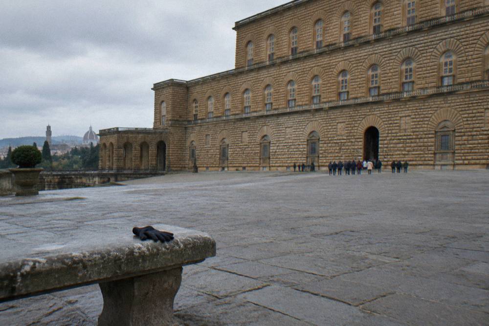 The wide sloping paving of Piazza de' Pitti before the vast rusticated facade of Palazzo Pitti, a loose line of visitors tiny and far off from behind near the palace doors