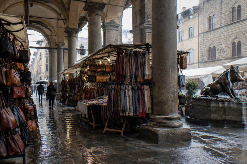 Inside the open stone loggia of the market, leather bags and scarves on trestle stalls under heavy vaulted bays, rain streaming off the arch edges, a scuffed stone pavement darkened wet