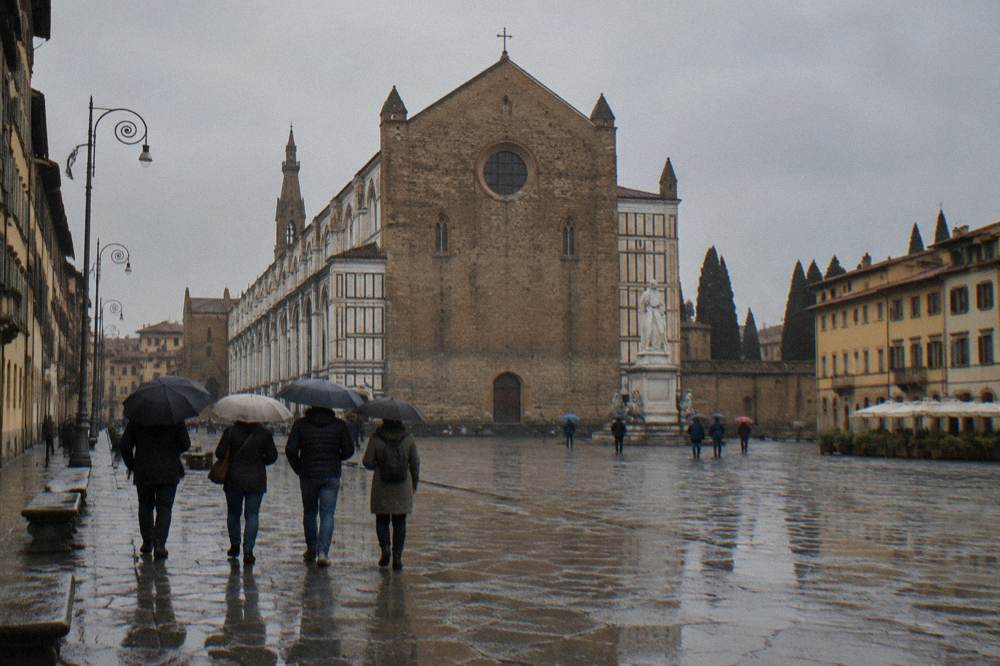 A small group with umbrellas seen from behind on the vast damp square, the church's rough stone gable and striped marble side panels ahead, puddles pooling between worn trachyte slabs