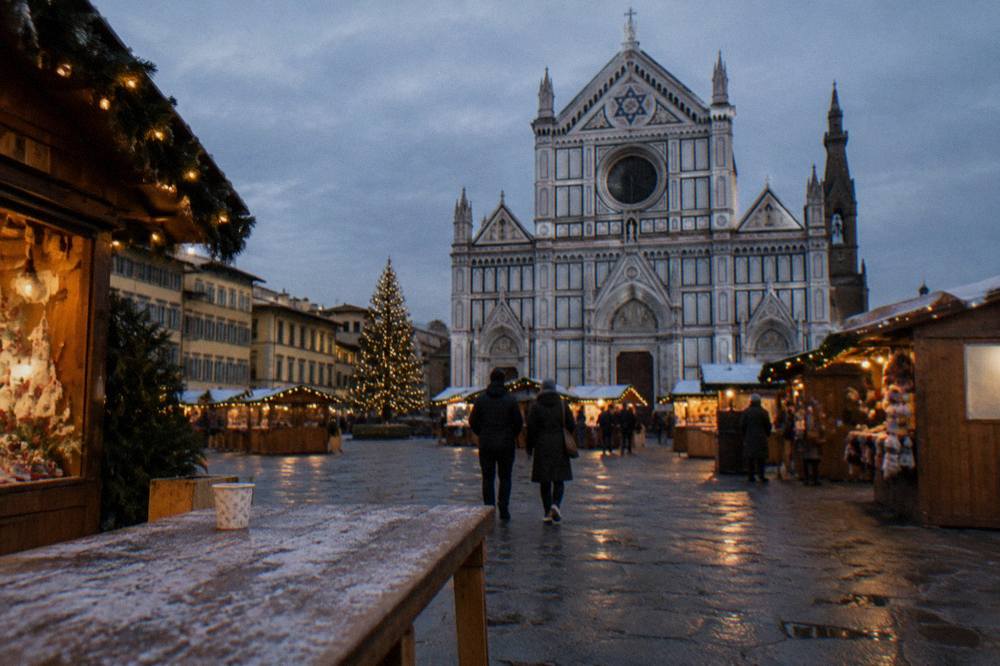 Wooden market huts ringing the vast stone square beneath the church's facade, warm bulbs strung along eaves, a paper cup left on a frost-slicked trestle