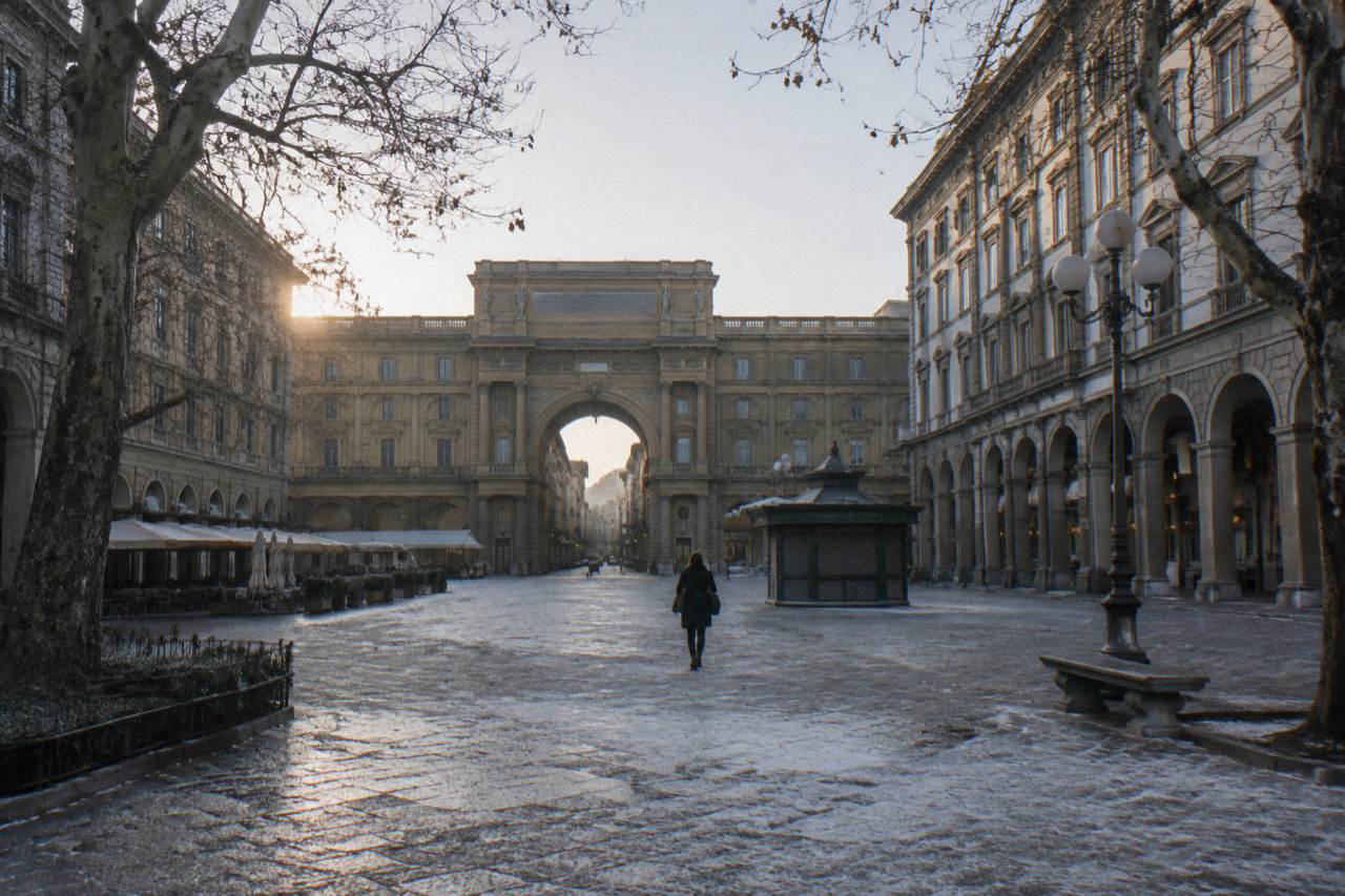 Looking down the long empty square flanked by tall plastered arcaded palazzi, bare plane trees at the edges, thin frost on the stone paving, one distant figure crossing from behind