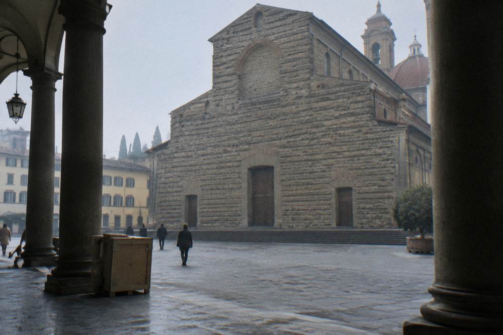 From inside the shadowed arcade across the square, looking at San Lorenzo's unfinished rough stone facade with its pilasters, a delivery crate propped by a column