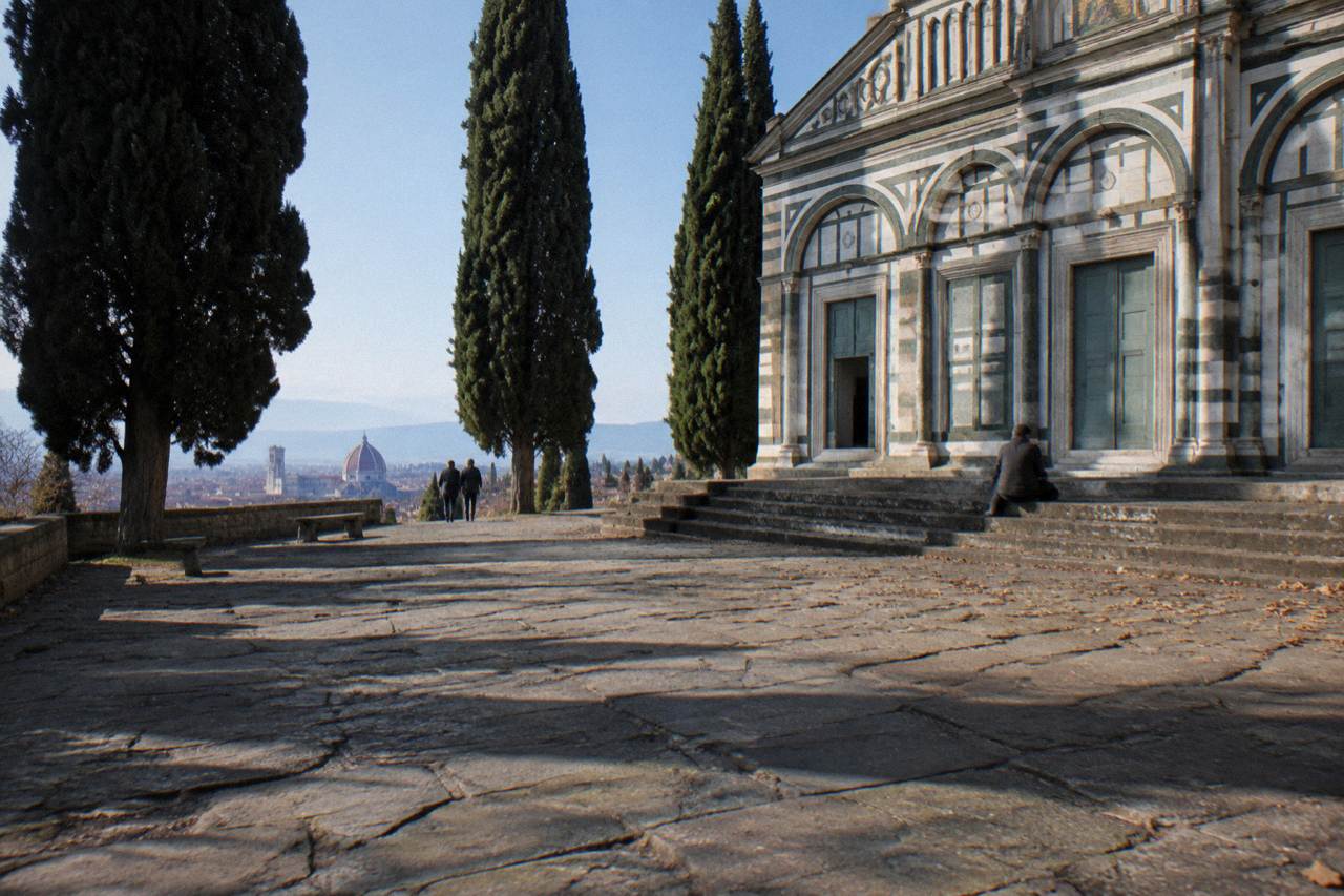 Low across worn churchyard flagstones toward the abbey's striped green-and-white marble facade, bare cypress shadows raking the pavement, a scatter of fallen leaves against the steps