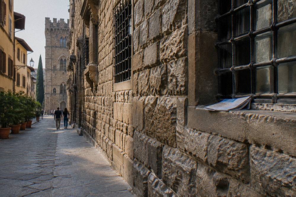 Looking along the side wall of the medieval Bargello palace from a narrow street, rugged stone blocks with barred windows set high, worn pavement, a folded paper map left on a window ledge