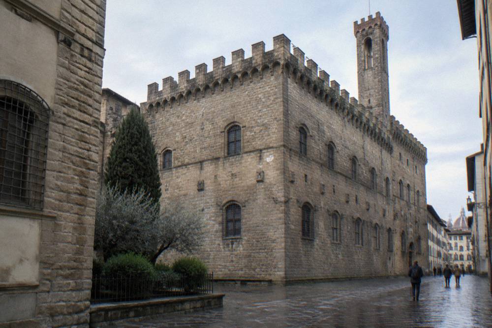 The rugged crenellated medieval stone corner of the Bargello palace on Via del Proconsolo, high pale stone walls with barred windows and a slender square tower above the roofline