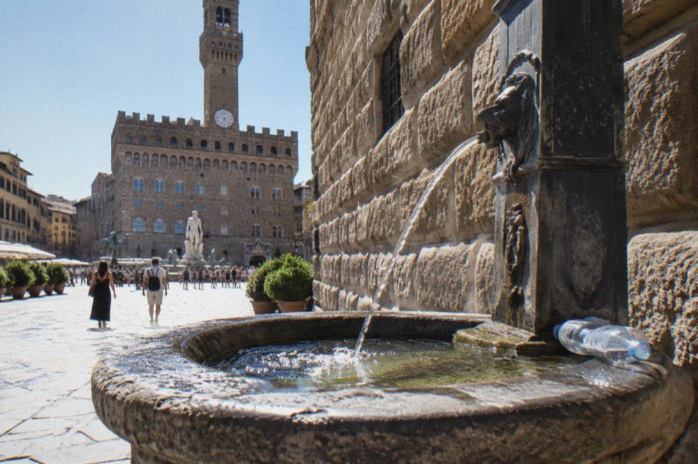 A public drinking fountain set against the rusticated pietra forte wall of Palazzo Vecchio, water arcing into a stone basin, sun glare on worn flagging