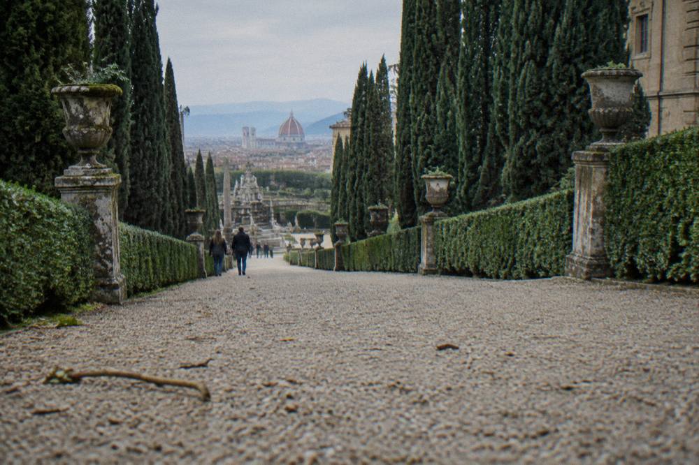 A formal avenue of tall cypress trees and clipped box hedges flanked by mossy stone urns, low camera close along the raked gravel, a fallen twig and scattered pebbles in the foreground
