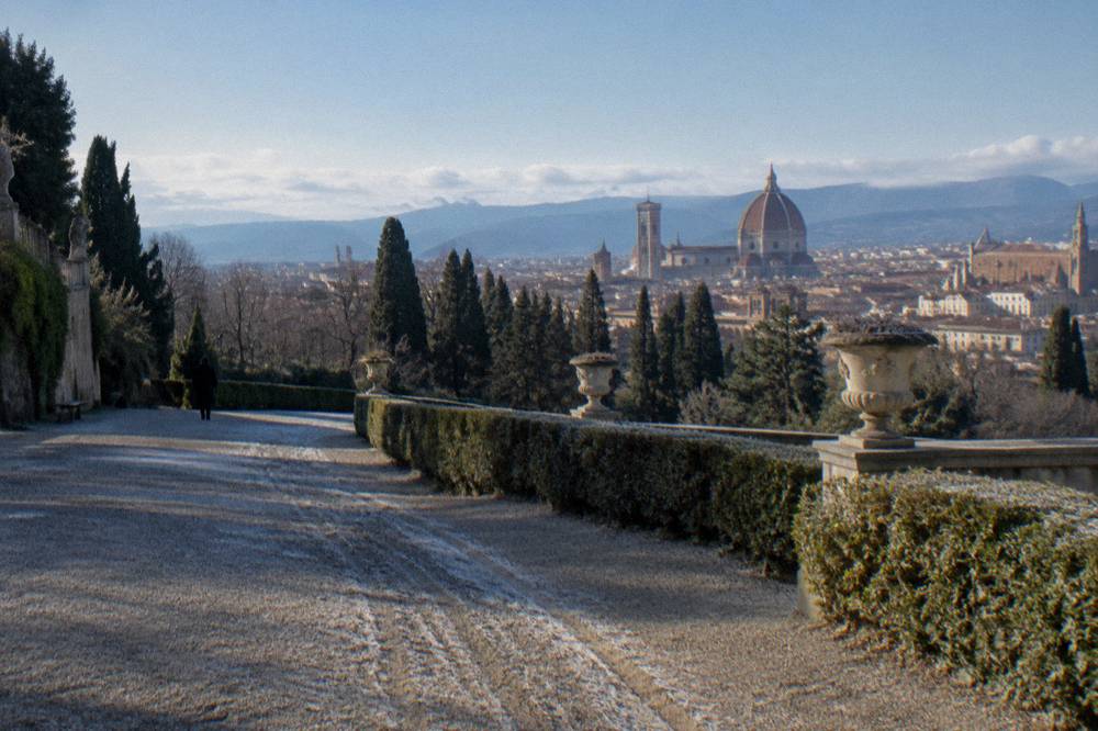 A high gravel terrace in the Boboli Gardens looking over bare cypress tops and terracotta rooftops to the Duomo, frost still in the ruts, a lone figure far off in a dark coat