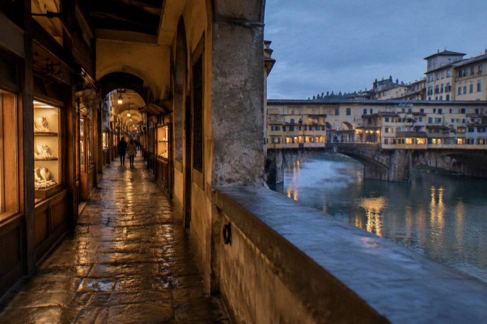 Looking along the enclosed stone passage of the Ponte Vecchio, small shop windows glowing gold onto worn flagstones, damp air after rain, a couple distant and out of focus