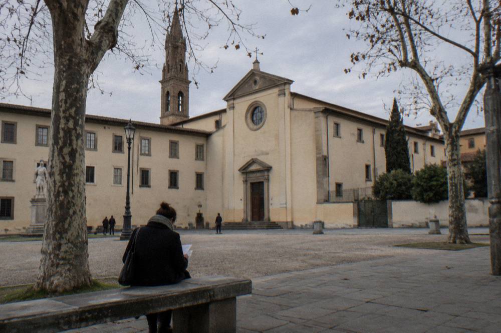 The plain pale facade of the church and convent on Piazza San Marco, bare plane trees, a stone bench, one visitor seen from behind reading a folded paper on the bench