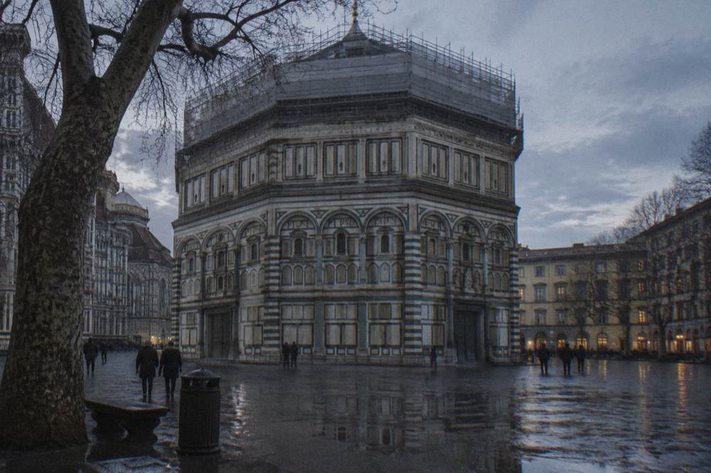 Striped marble walls below scaffolding wrapped along the roofline, bare plane trees and a puddled square reflecting the last grey light, Baptistery of San Giovanni