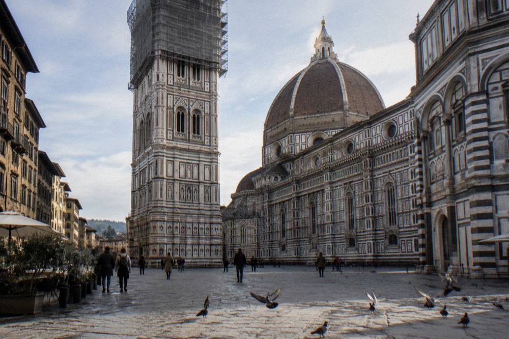 Giotto's striped marble bell tower beside the cathedral dome, scaffolding mesh and poles wrapped around its upper stages, pigeons scattering off the sunlit pietra forte paving