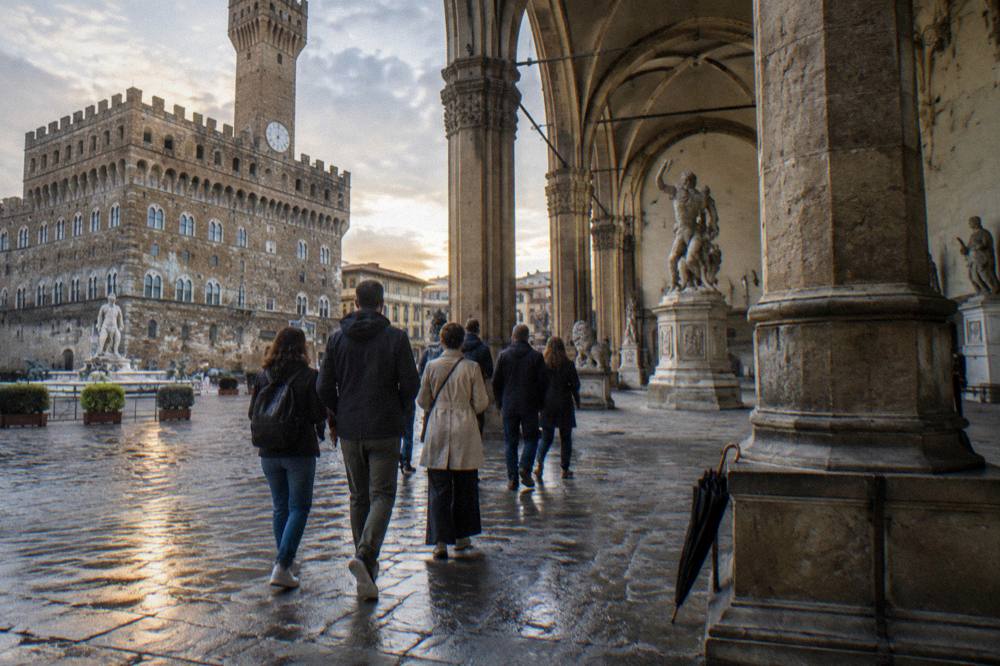 A small walking tour group seen from behind among the columns and arches of the Loggia dei Lanzi, slanting light on wet stone flagging, Piazza della Signoria