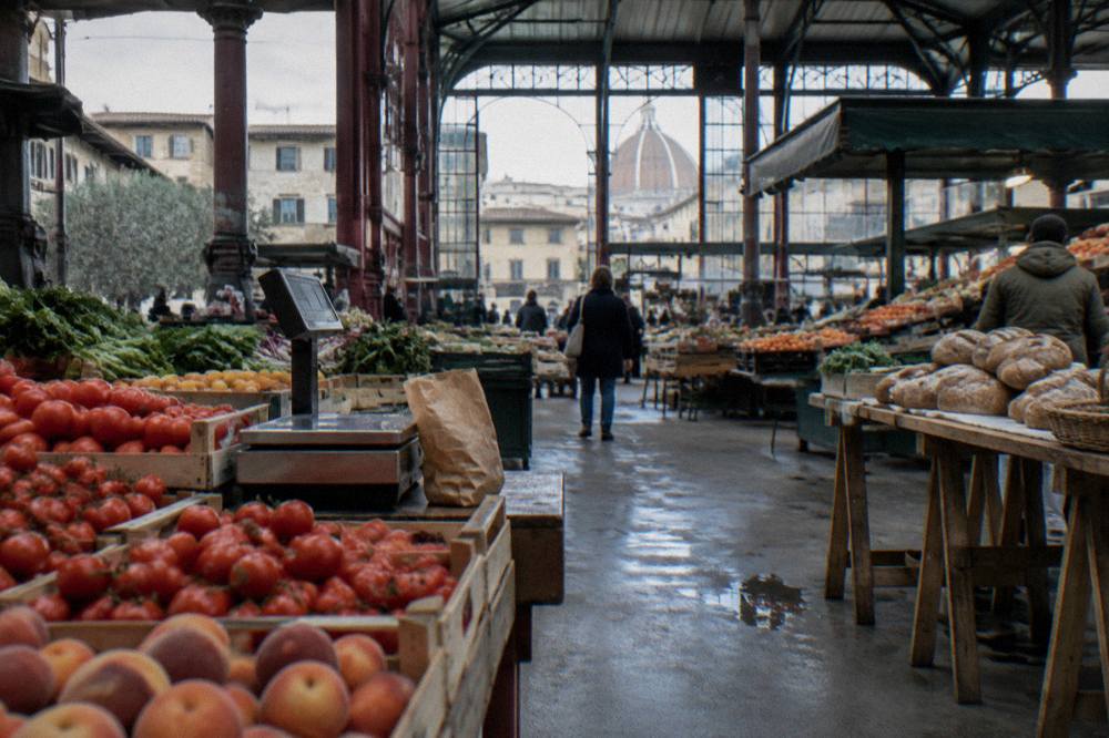 Inside the iron-and-glass market hall of Sant'Ambrogio, crates of tomatoes, peaches and round loaves on trestle tables, damp concrete floor, a half-empty paper bag left by a scale