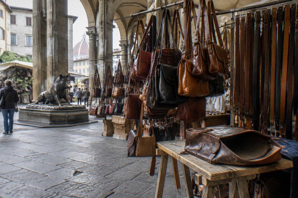 Under the long stone loggia of the Porcellino market, leather bags and belts hanging from stall rails, worn pietra forte arches behind, a scuffed leather satchel left open on a trestle