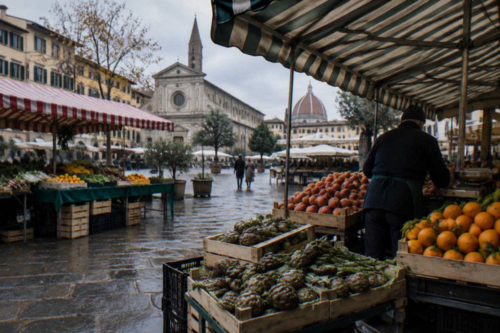 Produce stalls with striped awnings near the Sant'Ambrogio market, crates of artichokes and oranges, wet cobbles from a passing shower, a vendor seen from behind arranging peaches