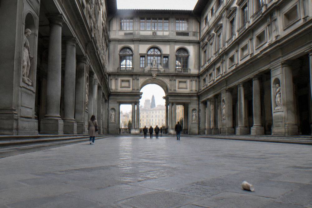 Seen up the long enclosed court of grey pietra sandstone arcades, a queue-less entrance, a dropped paper cup rolling on the smooth flagging under flat white light