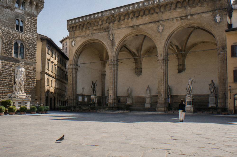 The open stone arches of the Loggia dei Lanzi on Piazza della Signoria seen from across the square, dark small shapes inside the arches, a pigeon on the pale paving