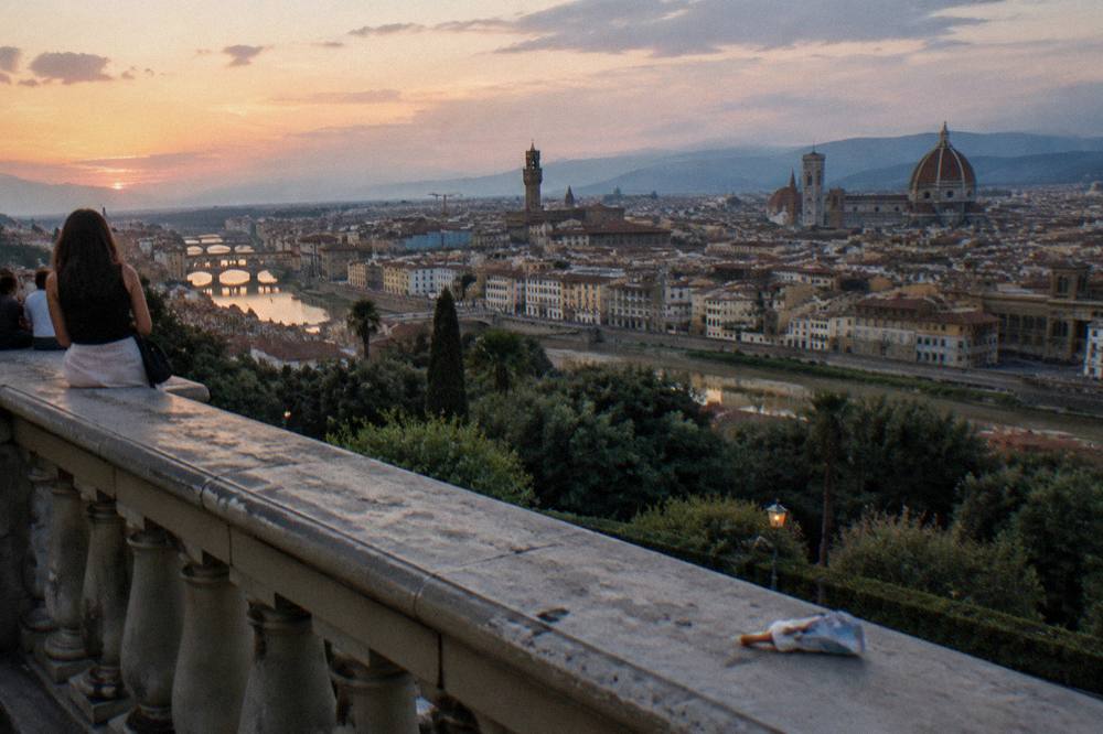 The Arno running low and green below, terracotta rooftops and the cathedral dome darkening, a dropped ice-cream wrapper caught against the balustrade, Piazzale Michelangelo