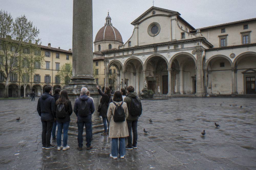 A small tour group gathered from behind around a guide beside the tall stone column, grey pietra arcades framing the square, wet pigeon droppings streaking the paving