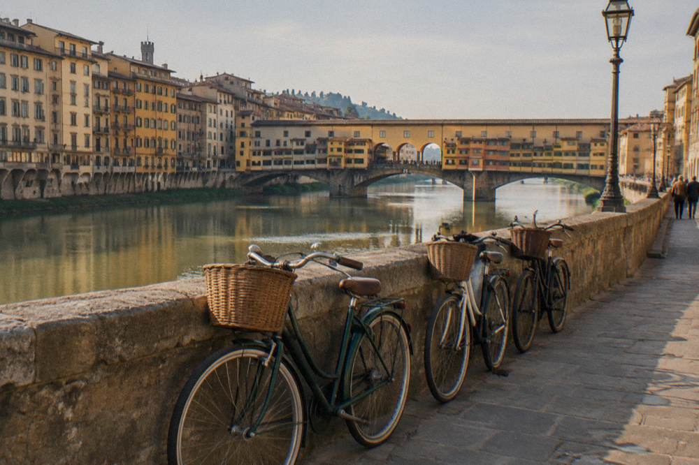 A row of old-fashioned bicycles with wicker baskets leaning against the stone parapet, the green Arno behind, no riders in frame, gold light on old stone