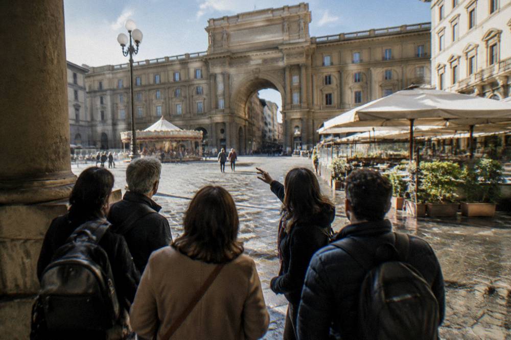 A small group seen from behind gathering at the arcade's edge, the guide's raised arm against the cafe awnings, wet cobbles drying in patches, Piazza della Repubblica