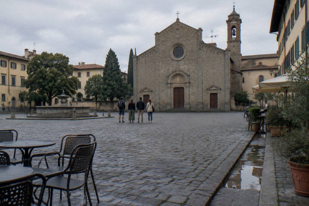 A small group standing far across the cobbles seen only from behind, facing the grey stone facade of the basilica, scattered cafe chairs and a puddled gutter nearby