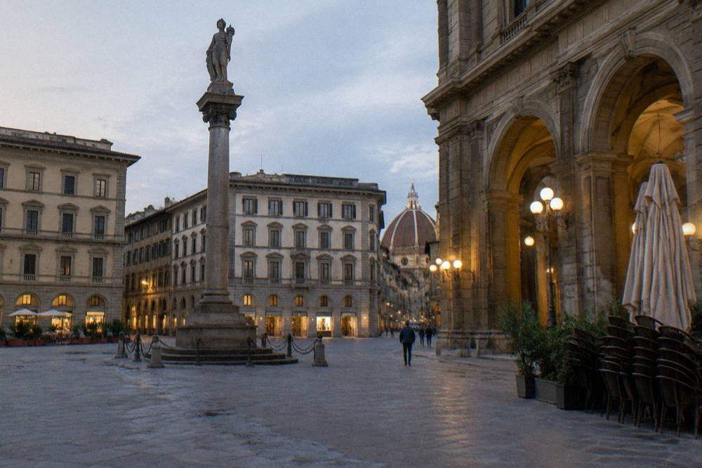 The tall stone Column of Abundance standing in the empty open square, cafe chairs stacked at the arcade edge, warm lamplight catching the grey pietra sandstone arches