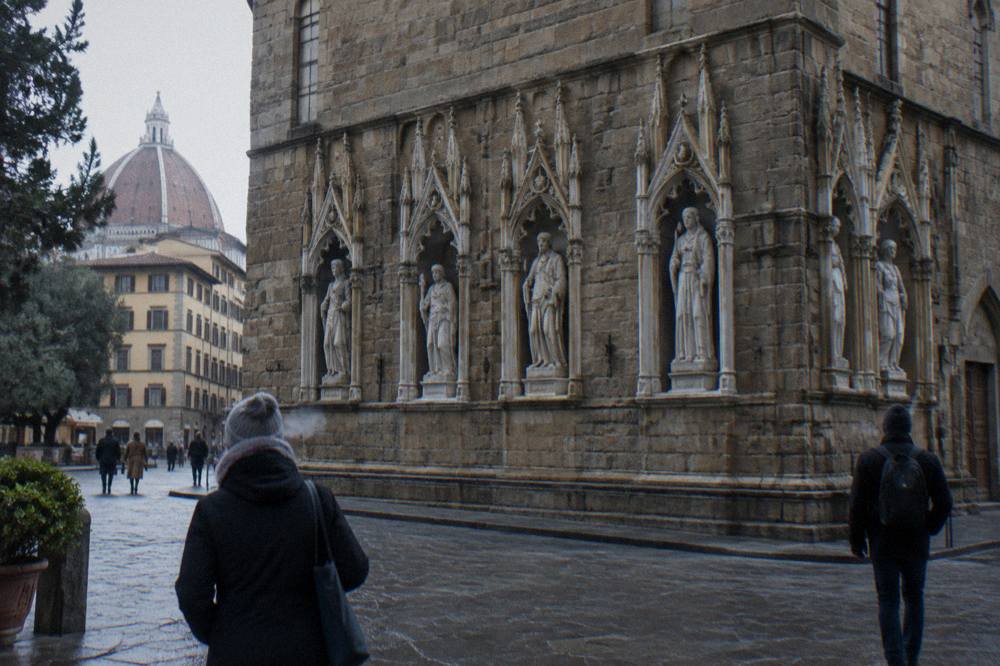Seen across the street toward Orsanmichele's rusticated stone facade, carved tabernacle niches running along its lower course, breath-fog and a couple of passersby in coats seen from behind