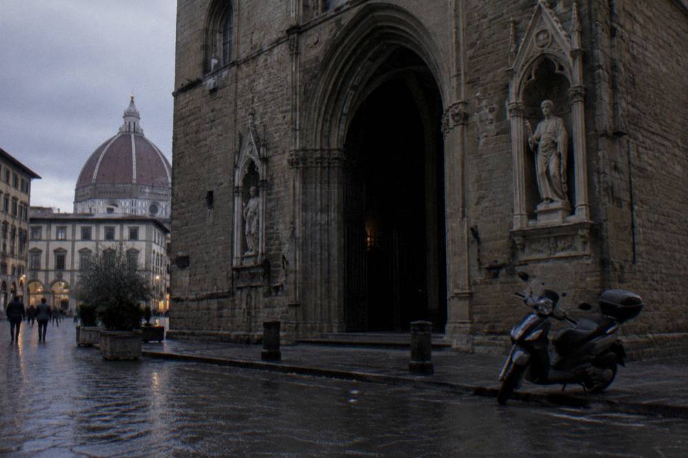 Seen across wet cobbles toward the shadowed arched doorway of Orsanmichele, its rough stone facade fading into grey, a scooter parked askew at the kerb