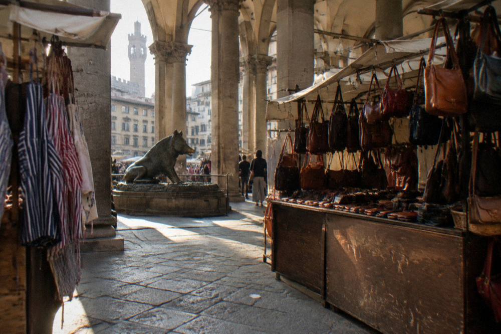 Stalls of leather bags and striped aprons in slanting light, a scuffed wooden stall counter, stray receipt paper on the flagstones, Mercato del Porcellino