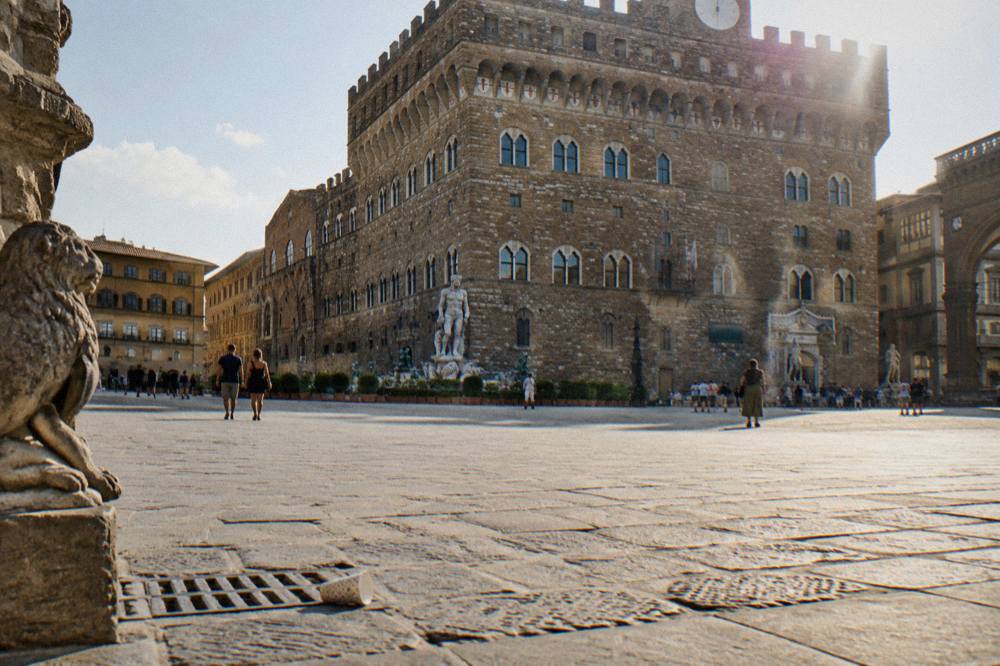 The arched windows of Palazzo Vecchio's stone facade seen across the sunlit paving of the square, a paper cup near a drain, a soft glare at the eaves, Piazza della Signoria